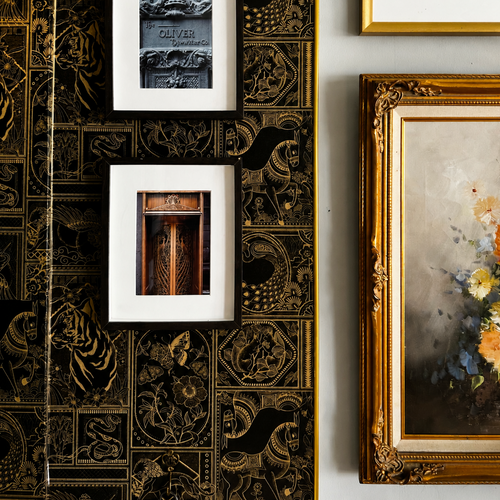 A styled interior wall featuring black and gold ornate patterned wallpaper, two small framed black-and-white photographs with white mats, and part of a larger gold-framed floral painting on a light gray wall.