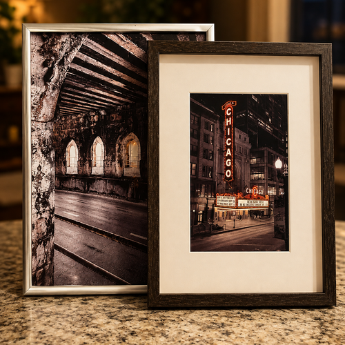 Two framed black-and-white Chicago cityscape photographs leaning against each other on a granite countertop. The left frame has a silver border and shows an arched stone passage; the right frame has a dark wood border, white mat, and a Chicago street scene with the Chicago Theatre sign.