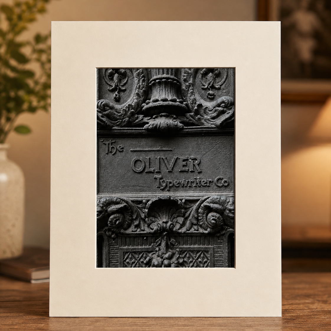 A framed black-and-white art print featuring an ornate cast iron sign reading 'Oliver Spawner Co,' displayed on a wooden surface with a blurred indoor background.