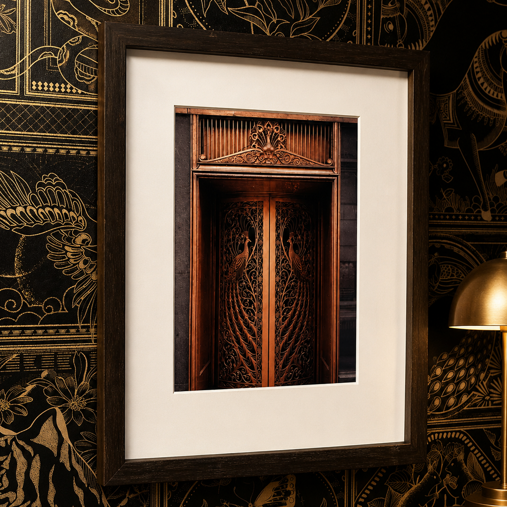 A framed art print featuring a detailed photograph of ornate wooden double doors, presented in a dark wood frame with a wide white mat, leaning against a black and gold patterned wallpaper background.
