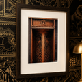 A framed art print featuring a detailed photograph of ornate wooden double doors, presented in a dark wood frame with a wide white mat, leaning against a black and gold patterned wallpaper background.