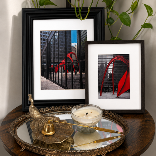 Two framed black-and-white urban art photographs with bold red arch sculptures, styled on a round wooden table with a mirrored tray, brass decor, and a candle. Green trailing plant leaves hang above the frames against a light neutral wall.