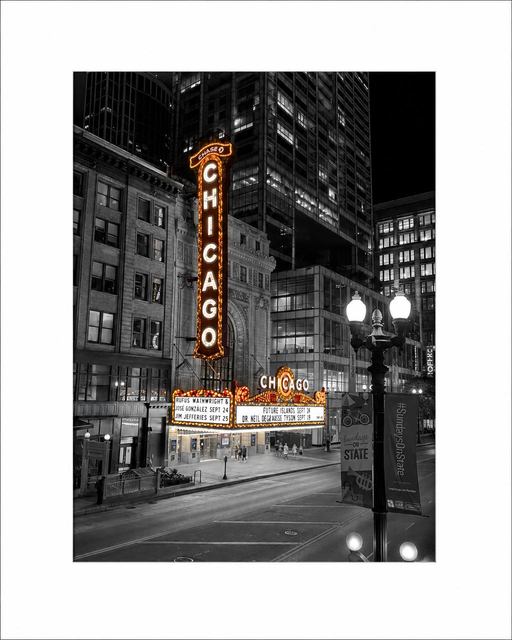 Black and white photograph of the Chicago Theatre at night with selective color highlighting the illuminated orange and yellow Chicago sign and marquee, surrounded by a white border.
